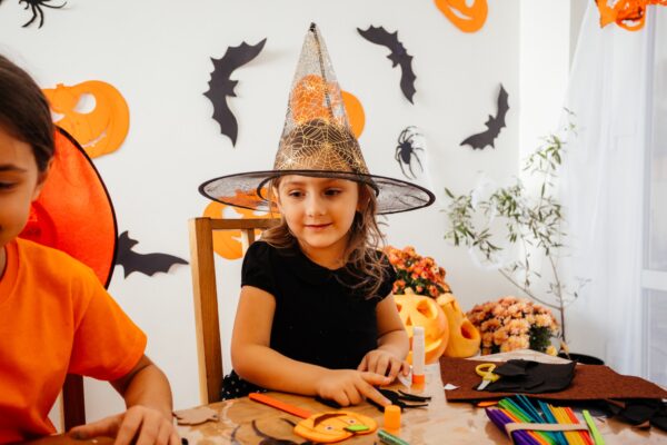 Adorable schoolgirls doing handmade paper decorations for autumn seasonal holidays. Engaged children working with paper and glue at wooden table. Halloween decor on white wall on a background.