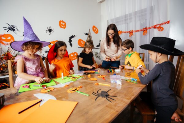 Cheerful and engaged schoolkids creating handmade bookmarks in halloween style during workshop. Teacher with children at wooden table making halloween decorations for school party.